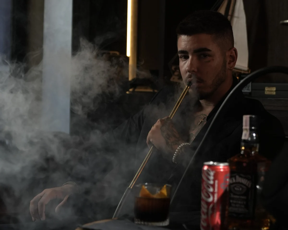 A man blowing a thick cloud of smoke while enjoying a premium hookah session with drinks, highlighting the bold flavors you find when searching for where to smoke & buy Mezza in Bali.