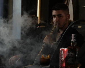 A man blowing a thick cloud of smoke while enjoying a premium hookah session with drinks, highlighting the bold flavors you find when searching for where to smoke & buy Mezza in Bali.