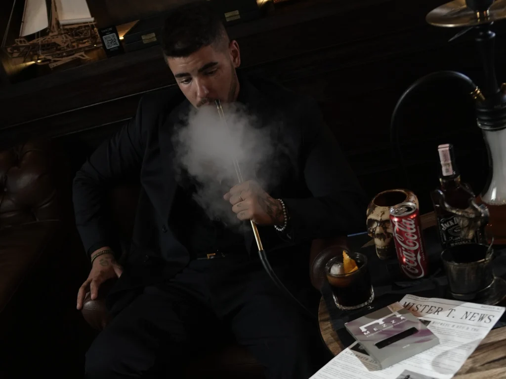 A sharply dressed man relaxing in a sophisticated lounge setting, exhaling a thick cloud of smoke while enjoying a smooth and flavorful dark leaf shisha session.