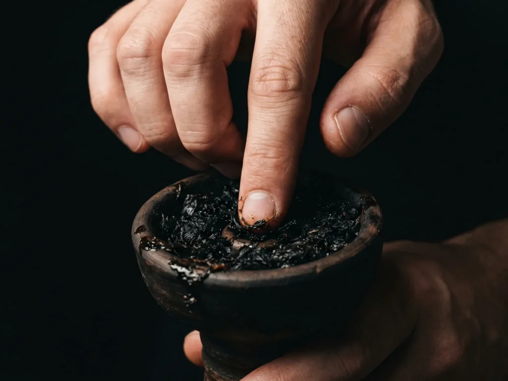 A close-up of a person's finger gently pressing down on the tobacco inside a clay bowl, demonstrating the proper dense packing technique required for a great dark leaf shisha session.